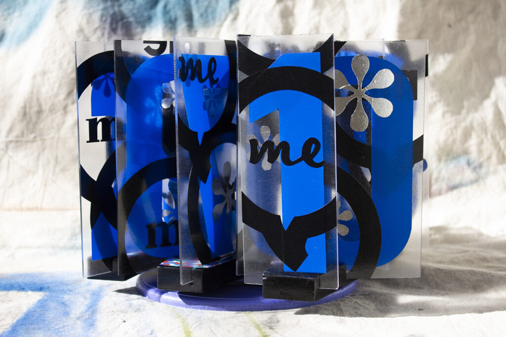 Sculpture of vertically placed acrylic letters each with a blue number, silver asterisk and black 'me' voice bubble on a piece of signage on a carousel