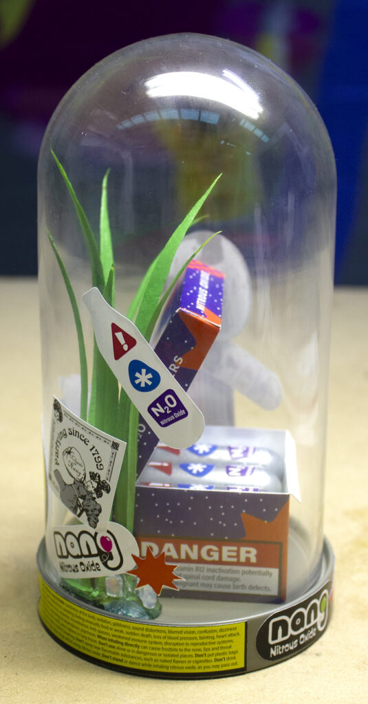 Sculpture of a fictional box of nitrous oxide chargers (Nangs) displayed amongst pebbles, grass and stickers