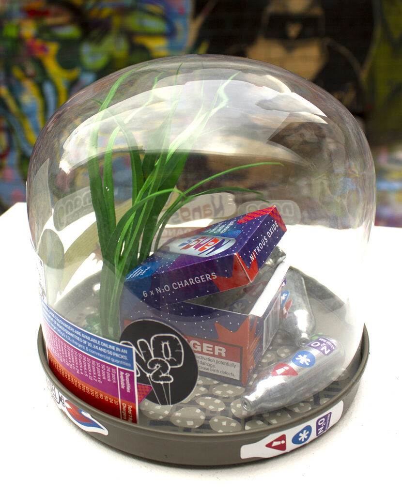 Sculpture of a fictional box of nitrous oxide chargers (Nangs) displayed amongst gravel, grass, empty chargers and stickers