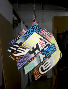 Hanging sculpture of an assemblage of aluminium composite signage letters with each side of the letter covered in a vignette of black to cream, then a coloured vignette pattern and various symbol characters